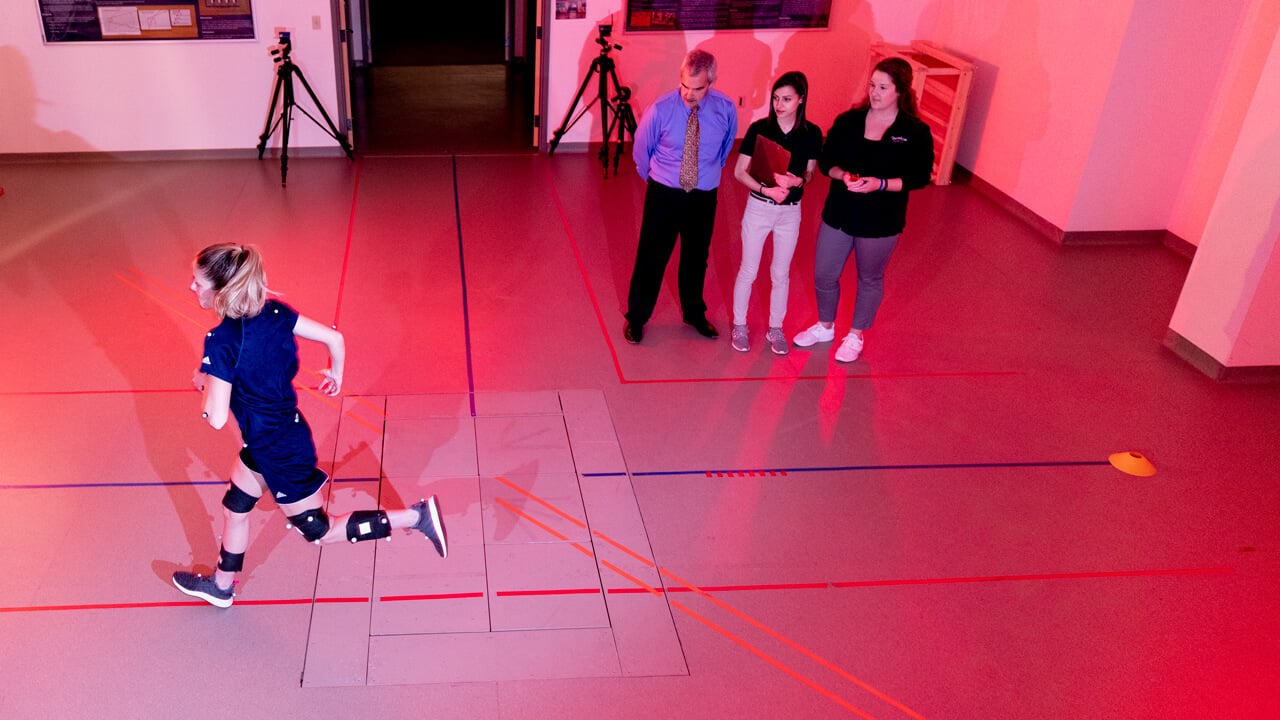 An athlete runs to the left in a motion analysis lab while a student and professor observe results.