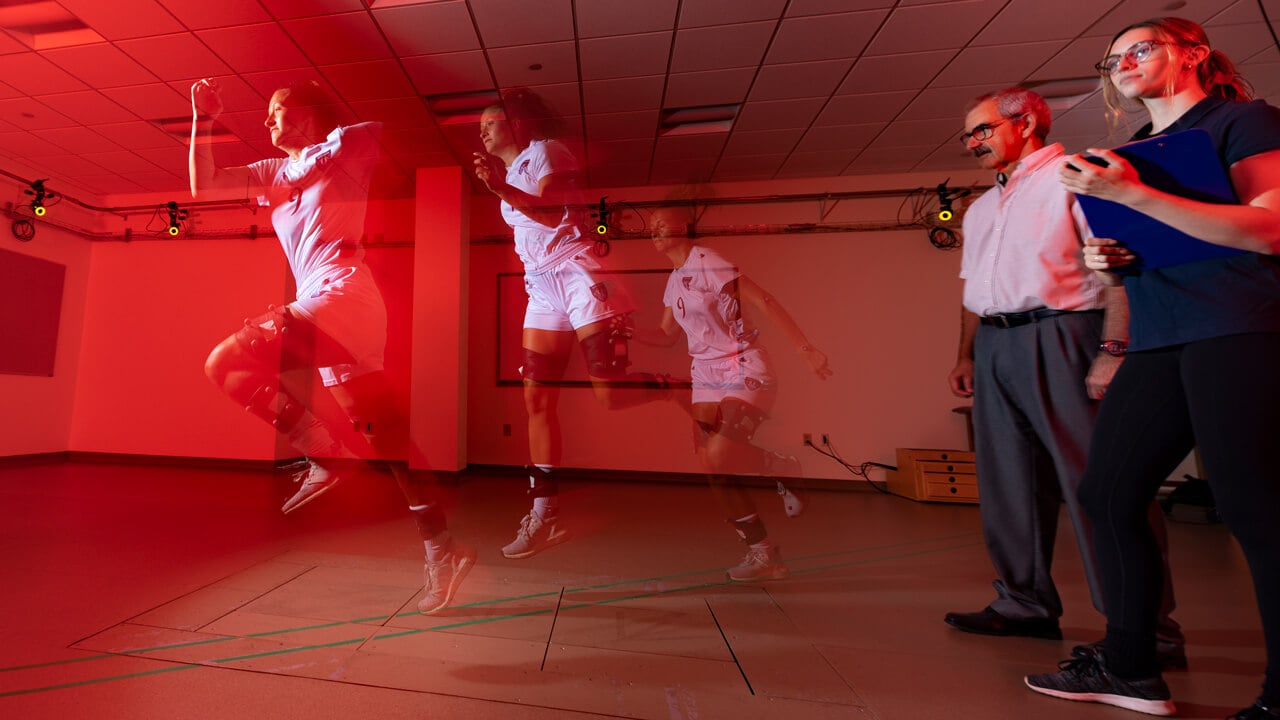 An athlete runs to the left in a motion analysis lab while a student and professor observe results.