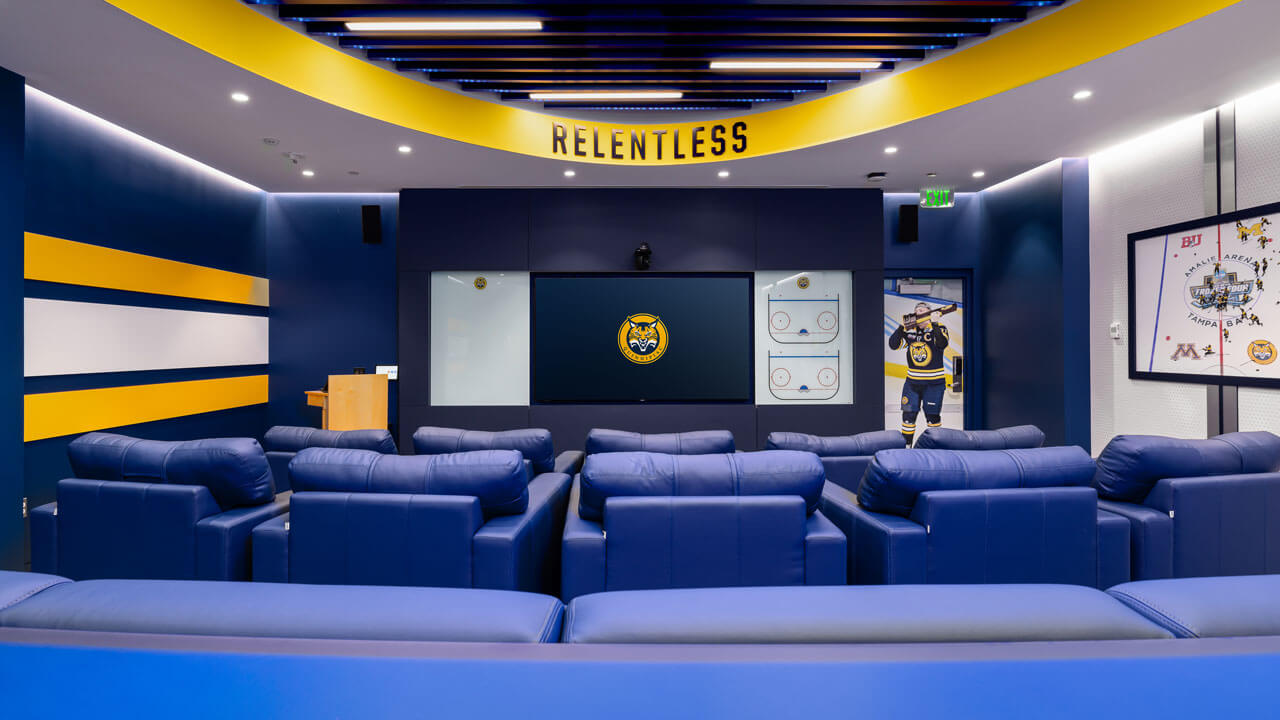 Wide shot behind the large blue chairs in the men's ice hockey locker room.