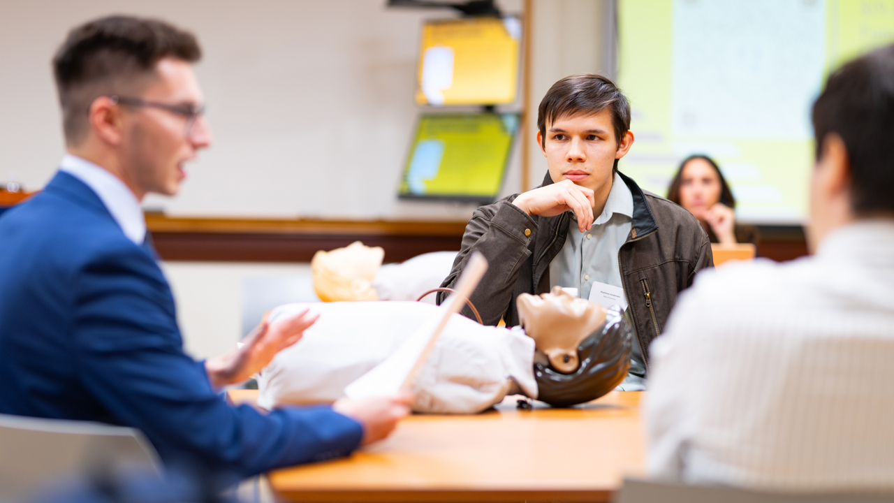 Three students converse with a CPR dummy between them.