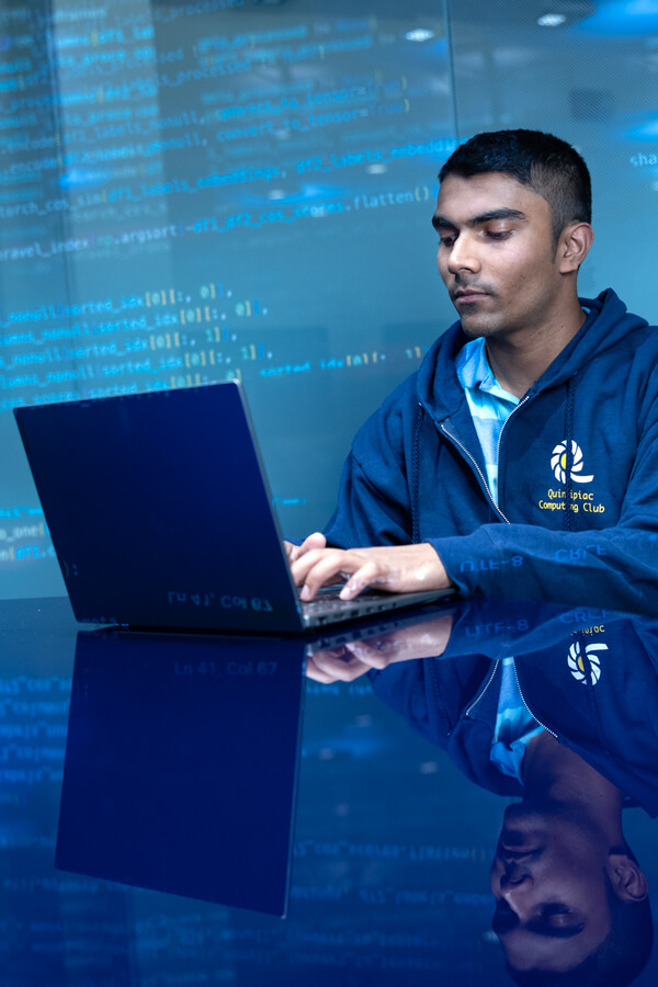 Student sitting down and working on a laptop with computer code projected on the back wall