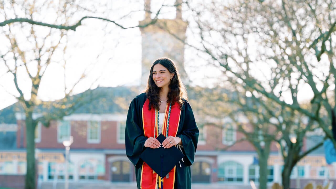 A student smiles wearing a graduation gown in front of the library.