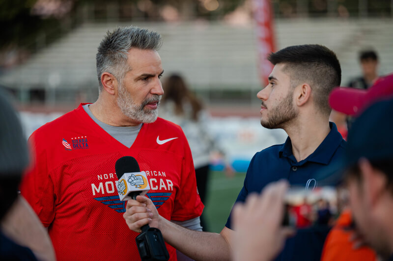 Santino Maione speaks with Kurt Warner on the sidelines of a football field