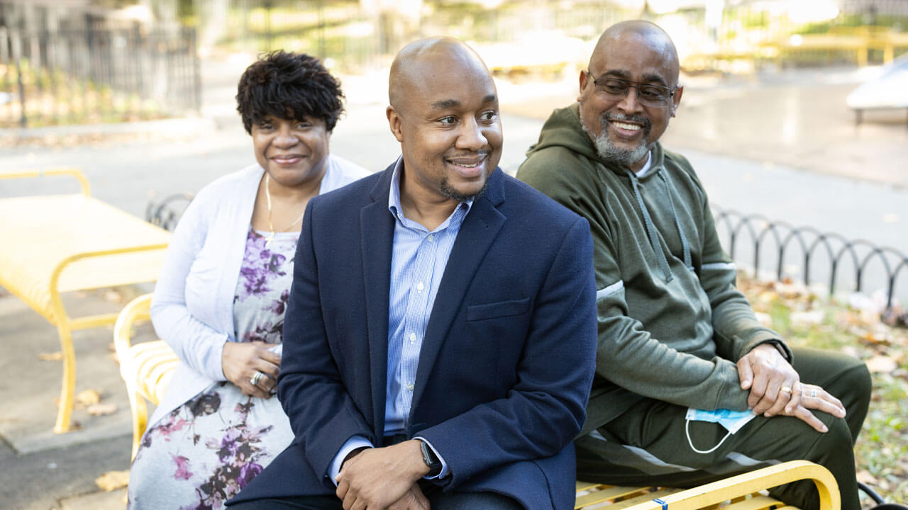 Don Sawyer sits with his parents on a bench outside in a Harlem neighborhood