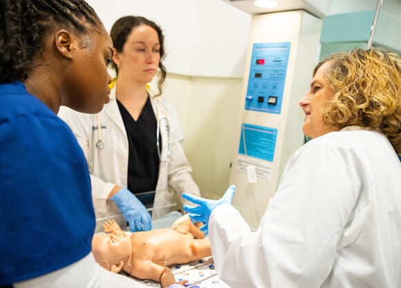 A professor instructs two nursing students over an infant mannikin