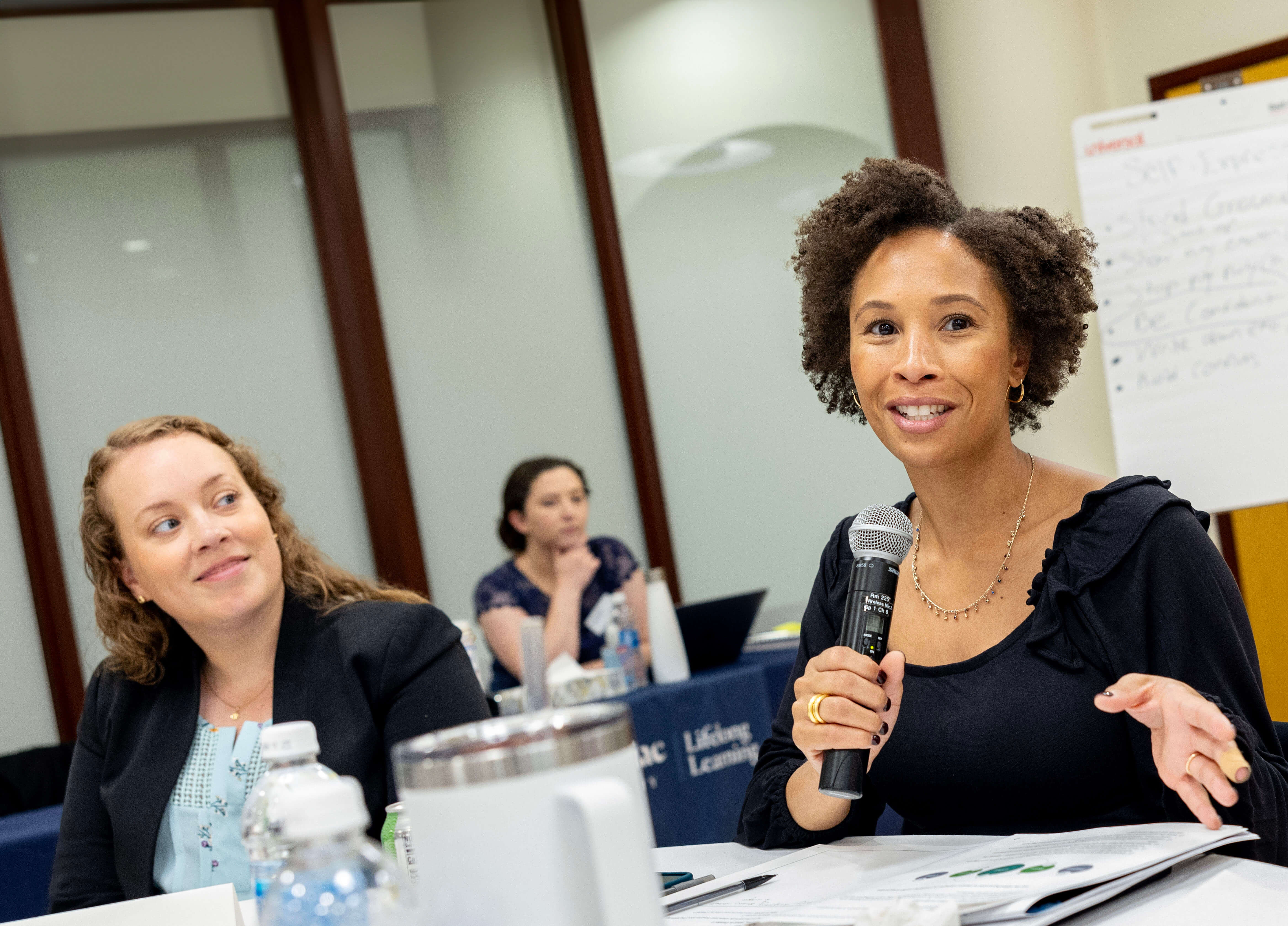 A woman in professional attire speaks into a microphone in a group setting.