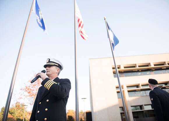 Jason Burke in his service uniform is holding a microphone speaking in front of an American flag