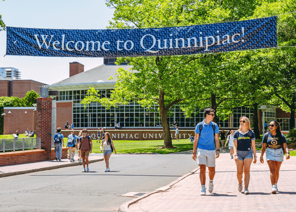Undergrad students walk and talk under a Welcome to Quinnipiac sign on Bobcat Way