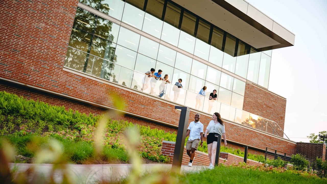 Students walk outside of the School of Business while others stand on the balcony.