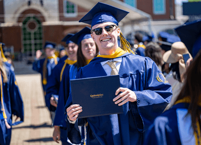 A graduate in sunglasses holds his diploma and smiles in front of the library clocktower