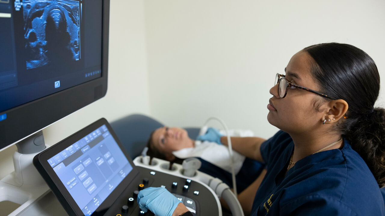 A female student conducting an ultrasound on another female's throat while using a control panel and screen