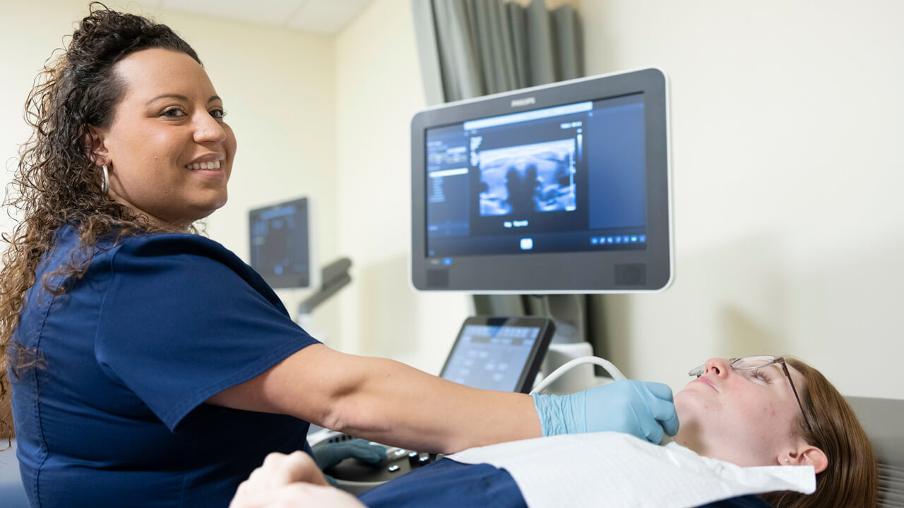 A female student conducting an ultrasound on another female's throat while looking off camera