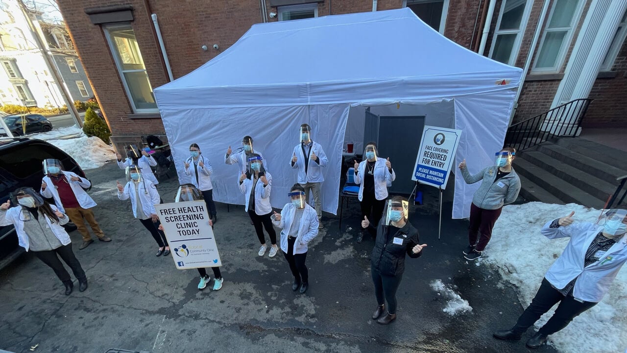 A shot from above of a group of students wearing masks and face shields and giving thumbs up outside a medical tent.