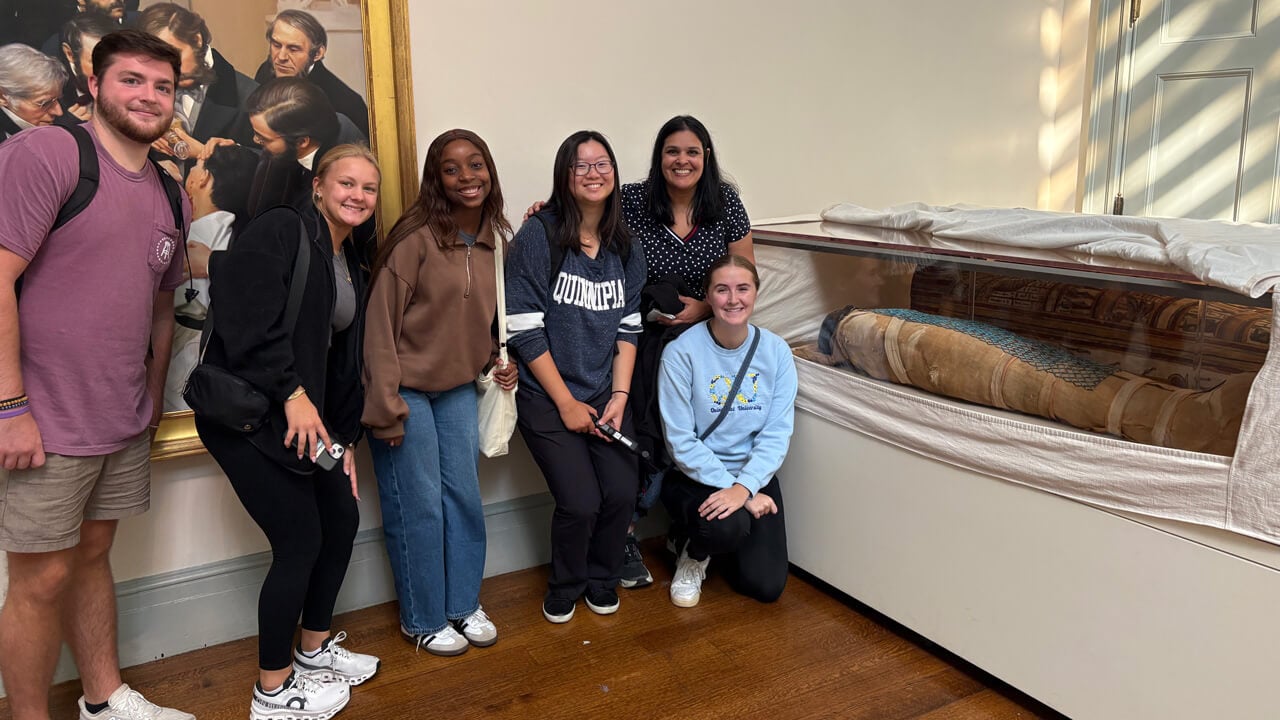 A group of six students stands and kneels next to a museum exhibit of a sarcophagus.