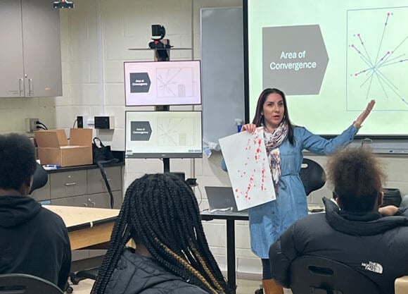 A professor teaches at the front of a classroom of high school students.