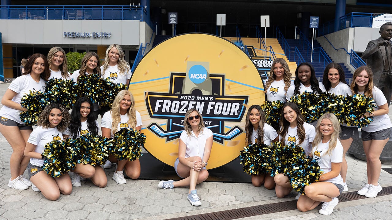 ice cats pose with their pom poms in front of puck shaped Frozen Four sign, their director sits on the ground in front