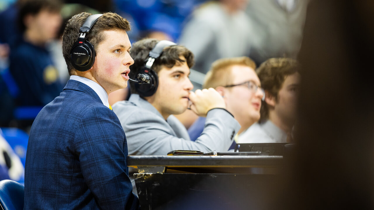 Three students sitting at a desk commentating on a men's basketball game