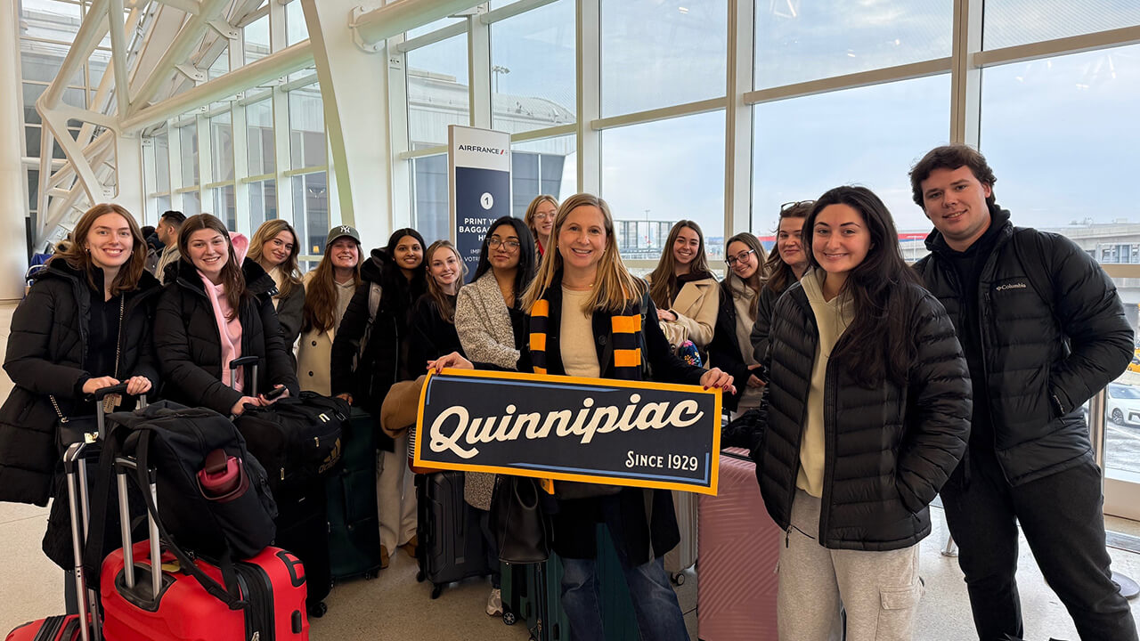 Students and faculty member gathered for a photo in the airport with a Quinnipiac sign
