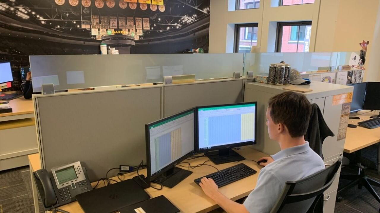 A male student sits at his desk in front of dual monitors working on spreadsheets