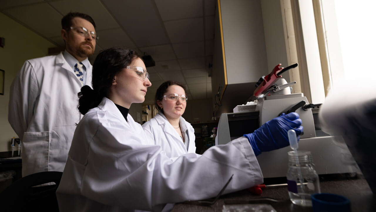 2 female student and a male professor work with a lab machine