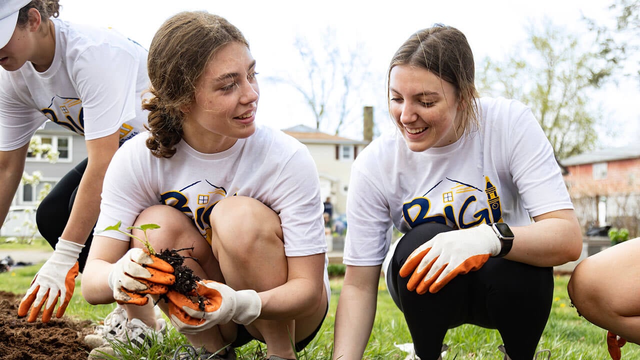 one female student holds up a small sprout, another female student digs a hole in the ground