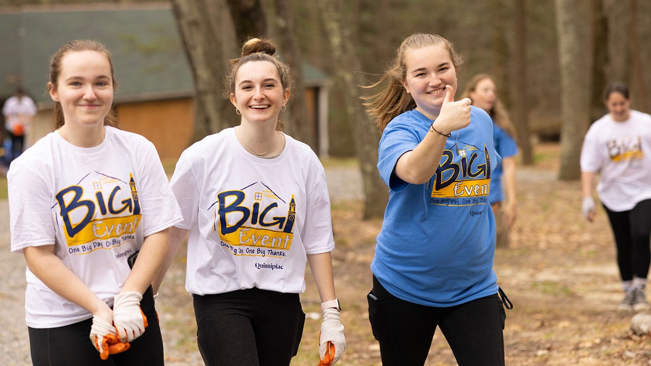 three smiling female students look towards the camera, one gives a thumbs up to the photographer