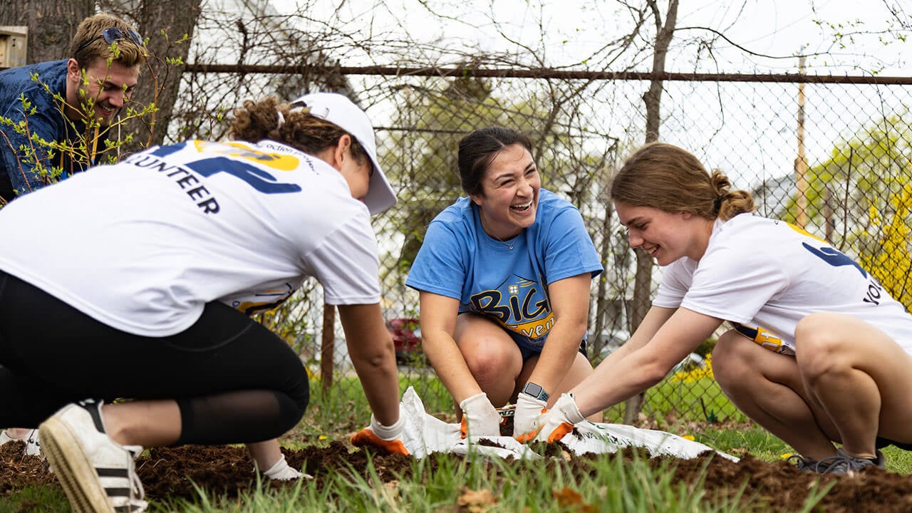 three female students open a bag of mulch, one looks to the side laughing at someone off camera