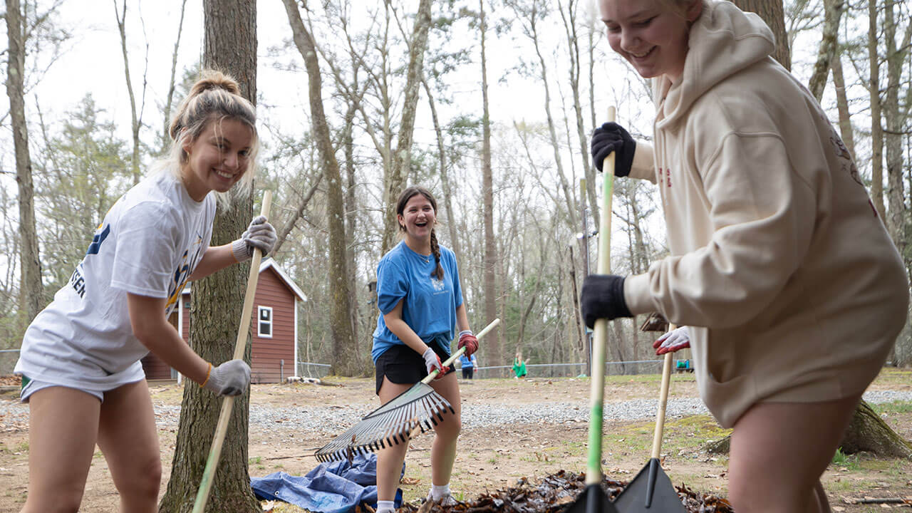 three smiling female students rake leaves onto a pile