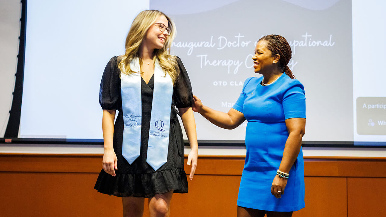 valerie sobol in a black dress and white graduation stole stands on stage for the occupational therapy ceremony with professor honoring her