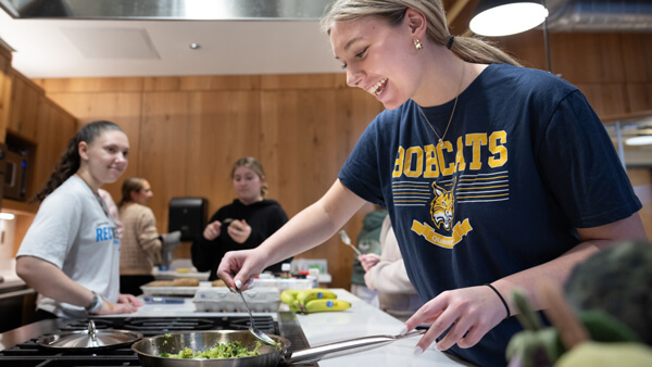 A student stirs broccoli in a silver pan in the teaching kitchen while other students look on