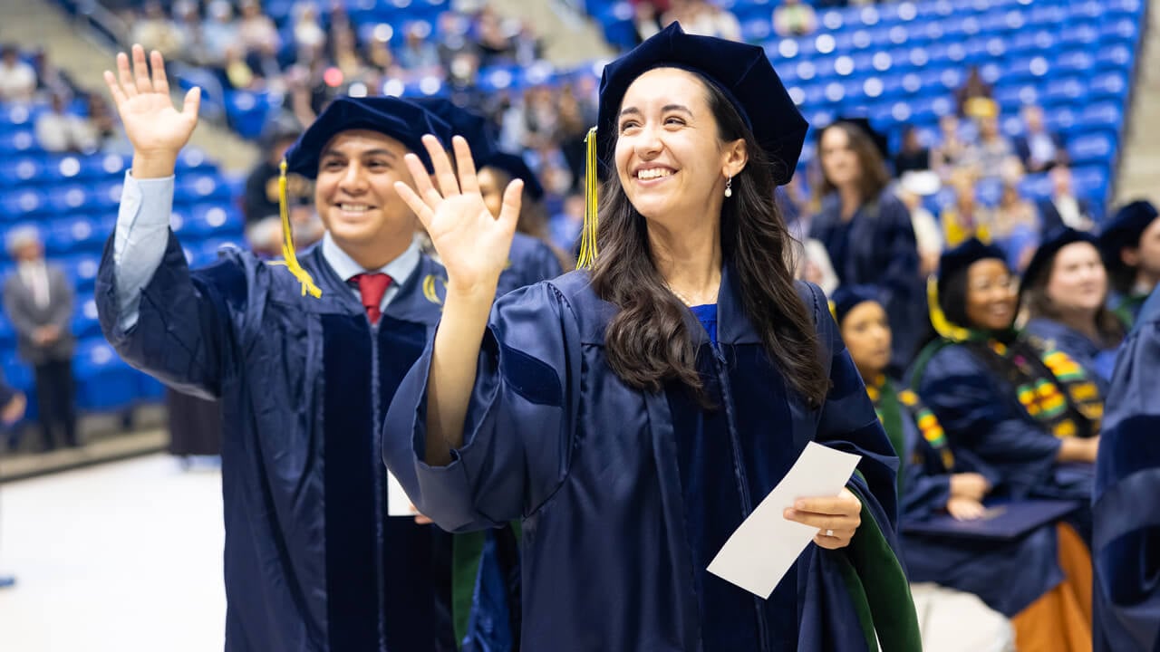 Two graduates smile and wave as they walk with their doctoral hoods draped on their arms