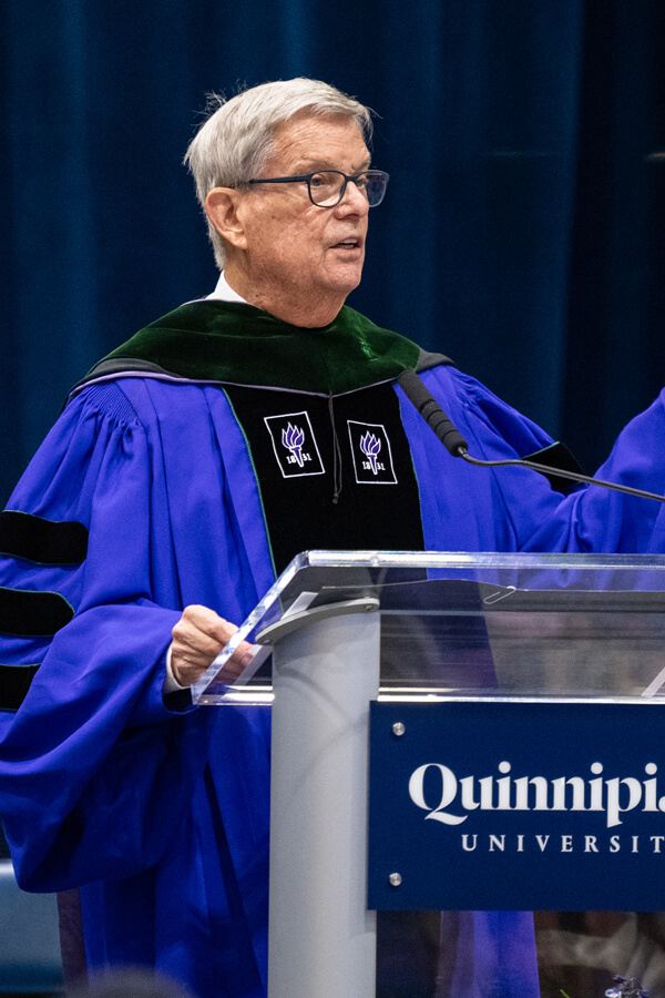 Richard Levin wears a cap and gown and speaks at a Quinnipiac podium at Commencement