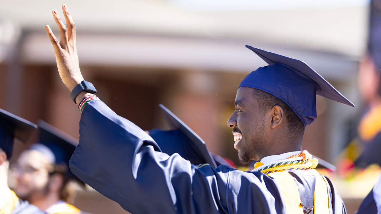 A graduate wearing green and gold cords waves