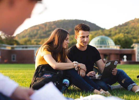 Students sit outside on the Mount Carmel Campus quad working on a laptop.