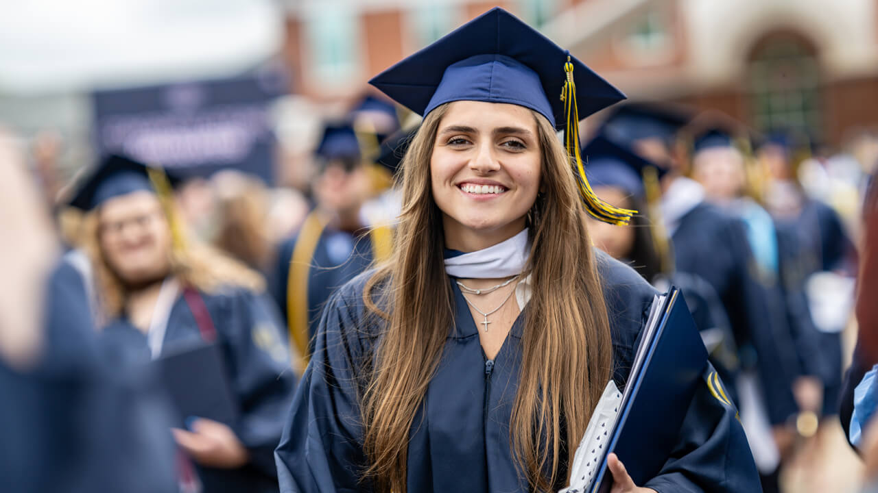 A graduate cradles her diploma and smiles as she walks in front of the library