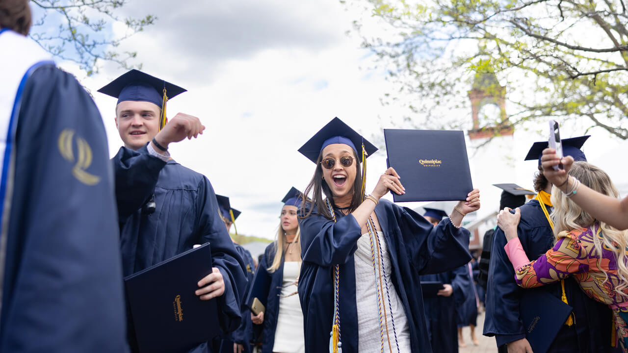 A graduate cheers and holds up her diploma in front of the clocktower