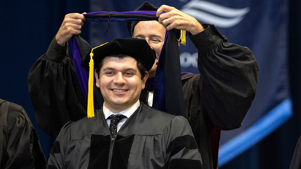 Graduate smiles as they receive their hood from faculty