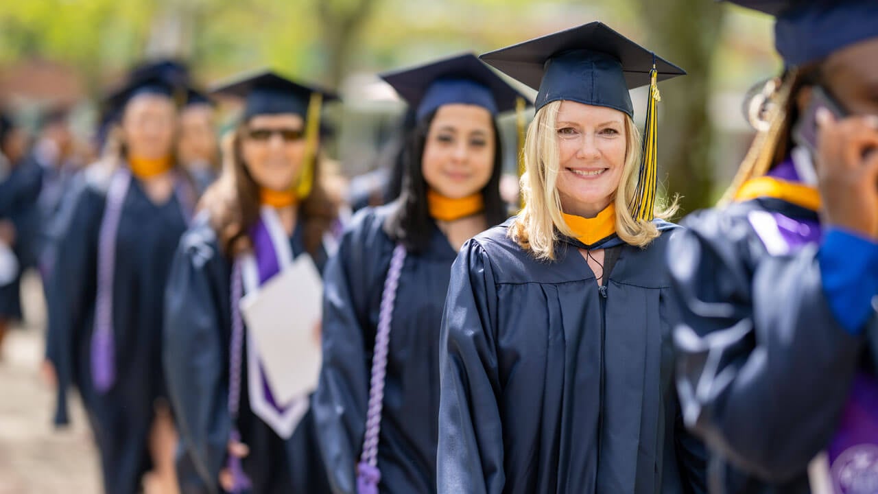 An elated class of 2024 graduate smiles brightly while standing in line amongst their peers.