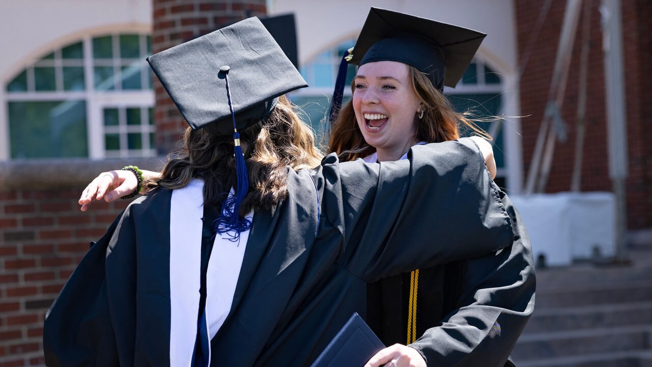 Two graduates exclaim and hug in front of the library