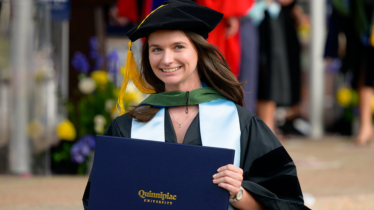 Graduate with teal stole and green hood smiles with diploma