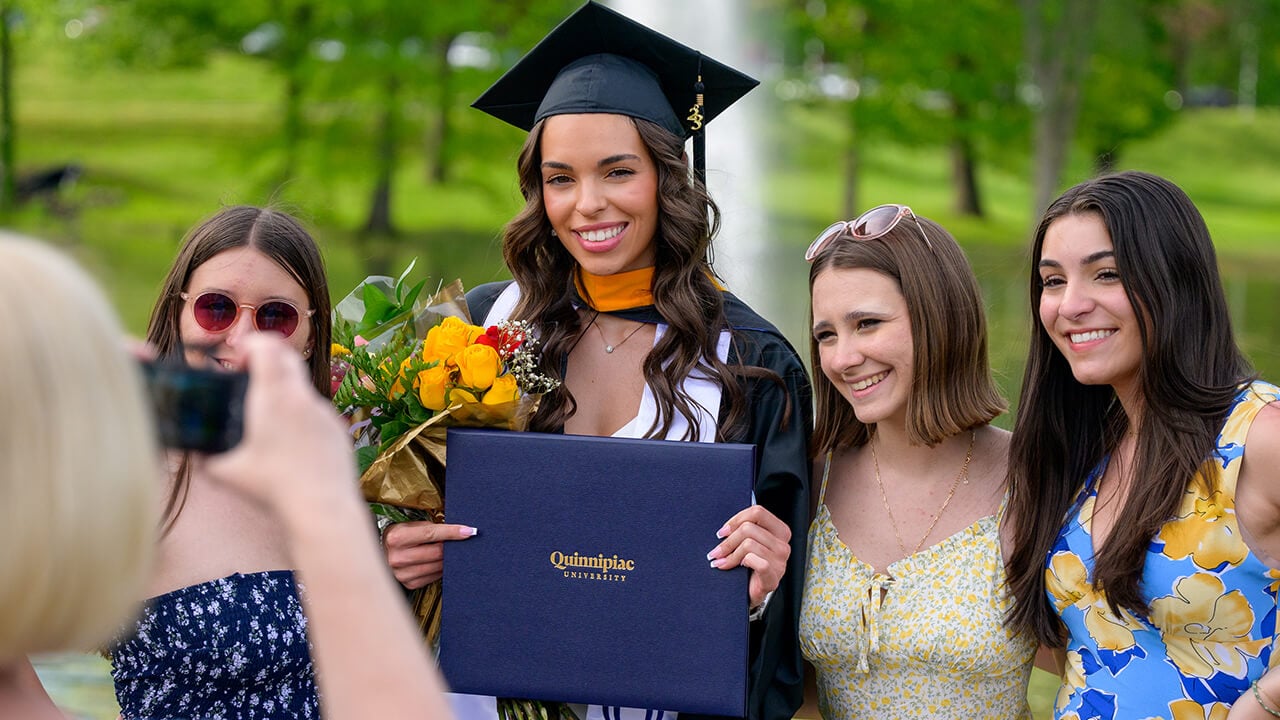 Graduate with flowers and diploma poses with friends in front of pond fountain