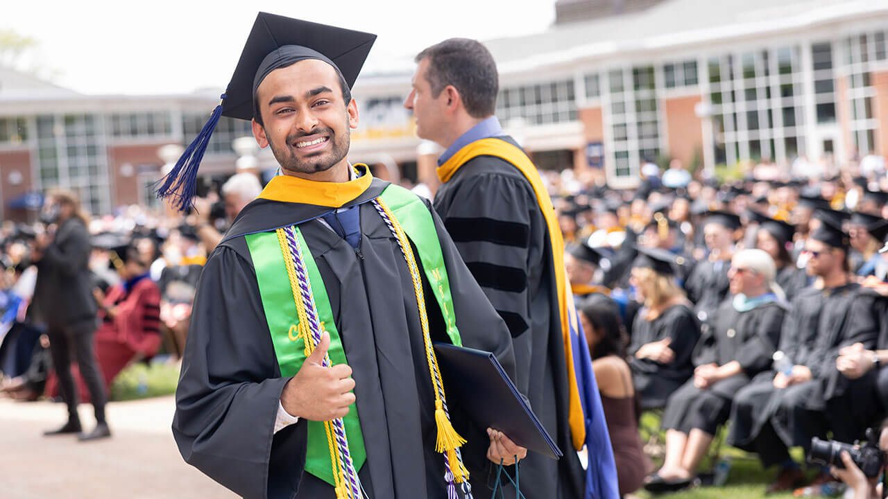 Student with a cap and gown on holding a thumbs up in front of the student center