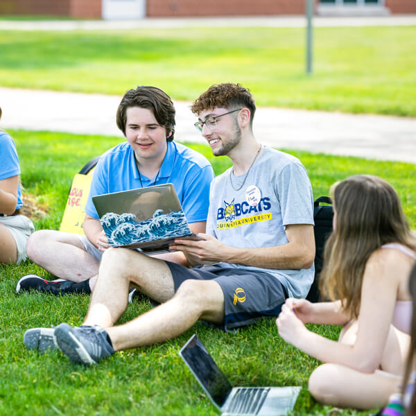 Two students sit on the grass at Orientation and look at a laptop with blue waves on it