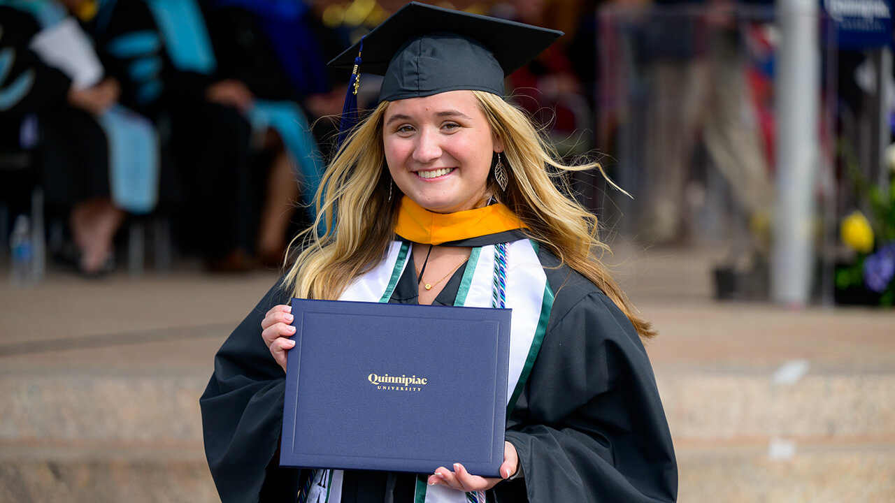 Graduate adorned with cords and a stole smiles posing with diploma