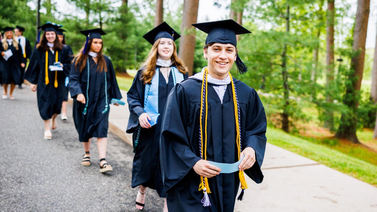 Graduates walk through a row of trees headed to the quad