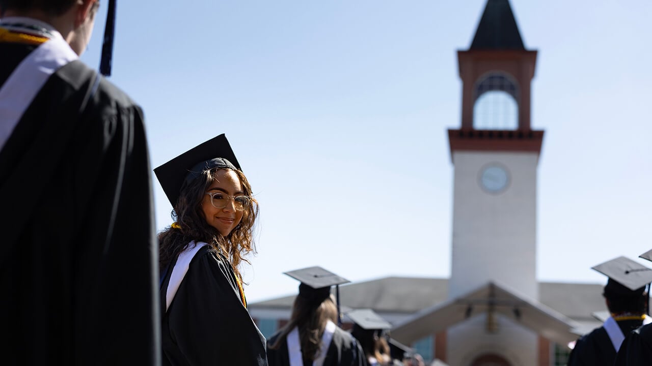 Graduate turns around to camera whilst walking in the processional in front of Arnold Bernhard Library