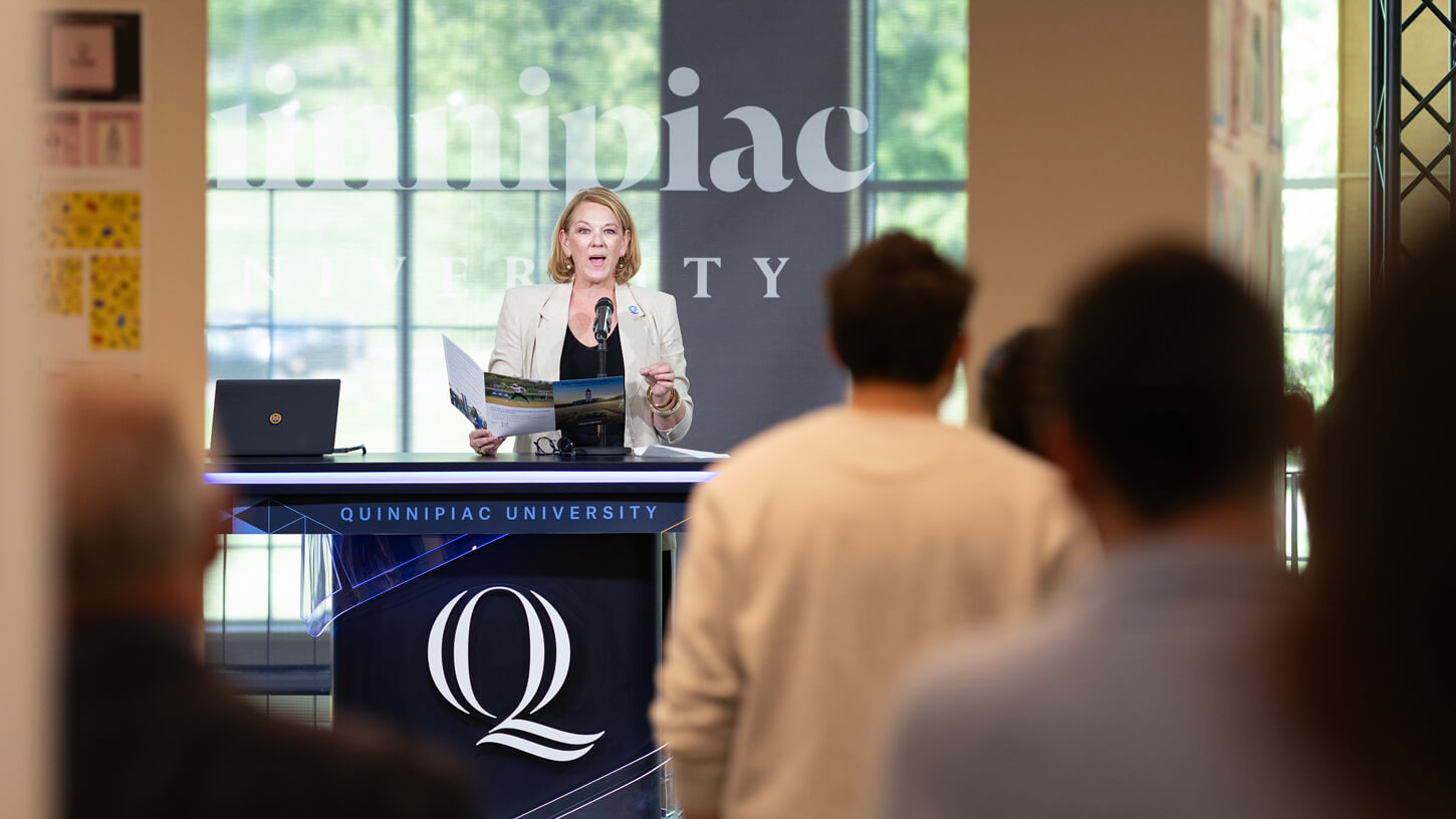 A female speaker presents to an audience at a Quinnipiac University-branded podium, holding a program booklet.