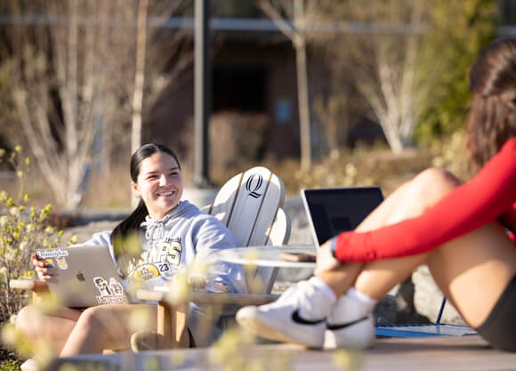 Two students sit in Adirondack chairs and talk on a spring day