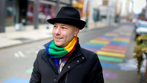 Vince Contrucci walks down a street painted in rainbow colors wearing a rainbow scarf.