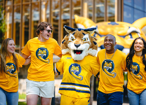 Four students and the bobcat mascot lock arms and walk together wearing matching shirts